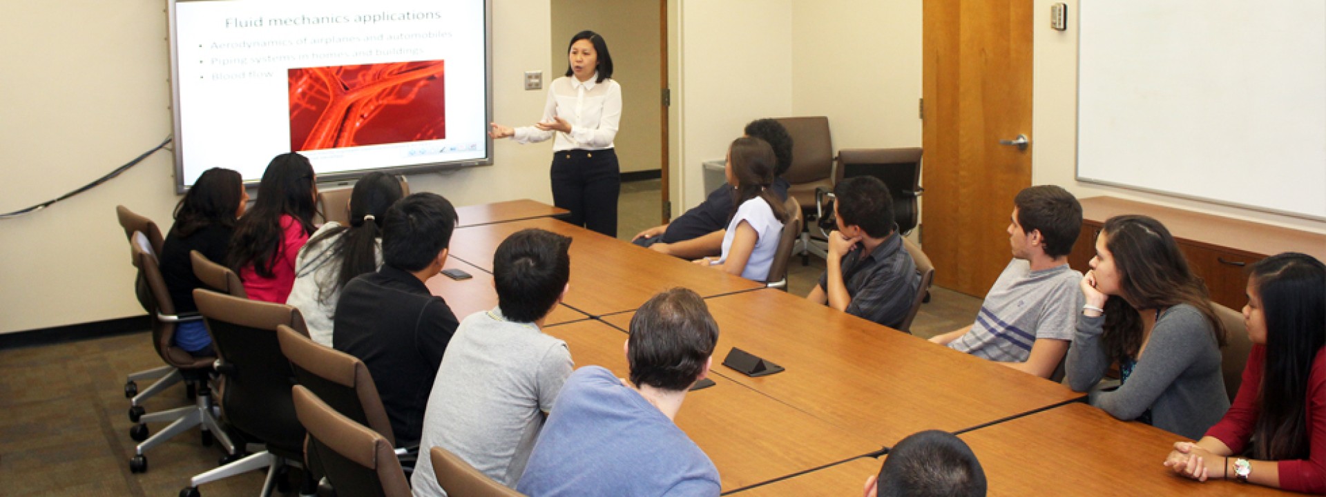 Group of Cal State LA students in a classroom around a conference table facing a projector screen as a lecturer teaches.