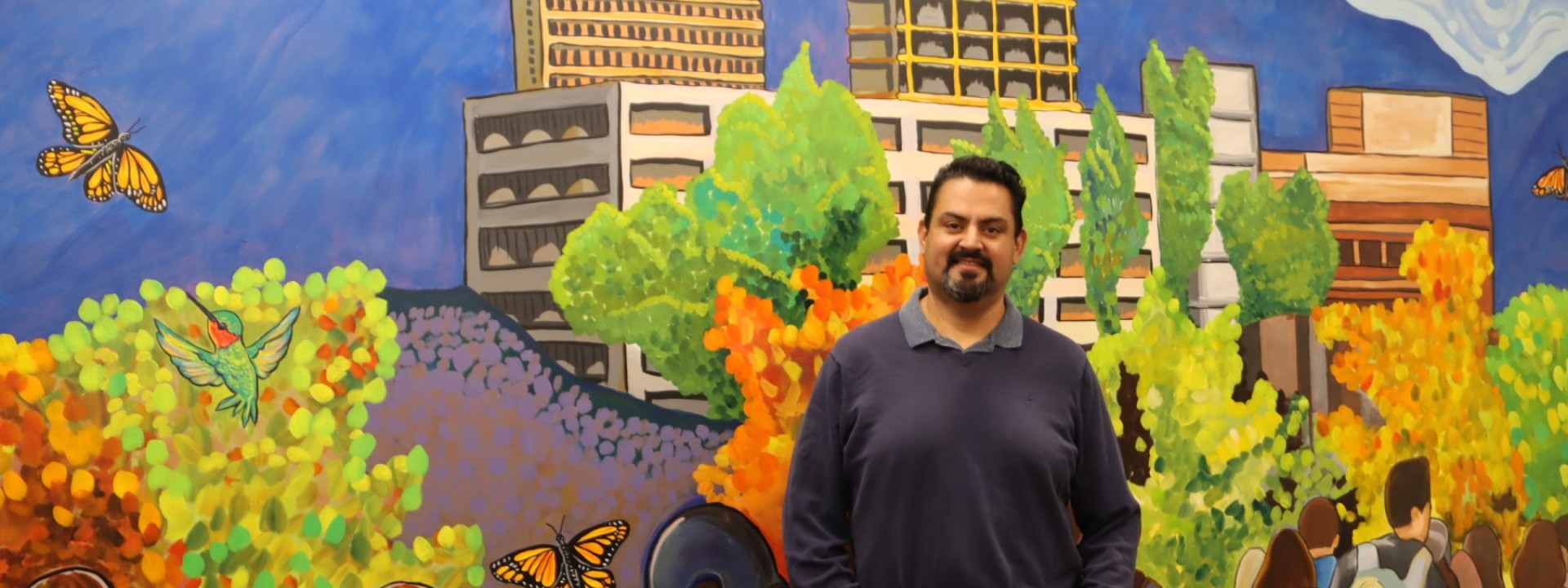 MA Student, Reynaldo Mora posing in front of a mural he painted 