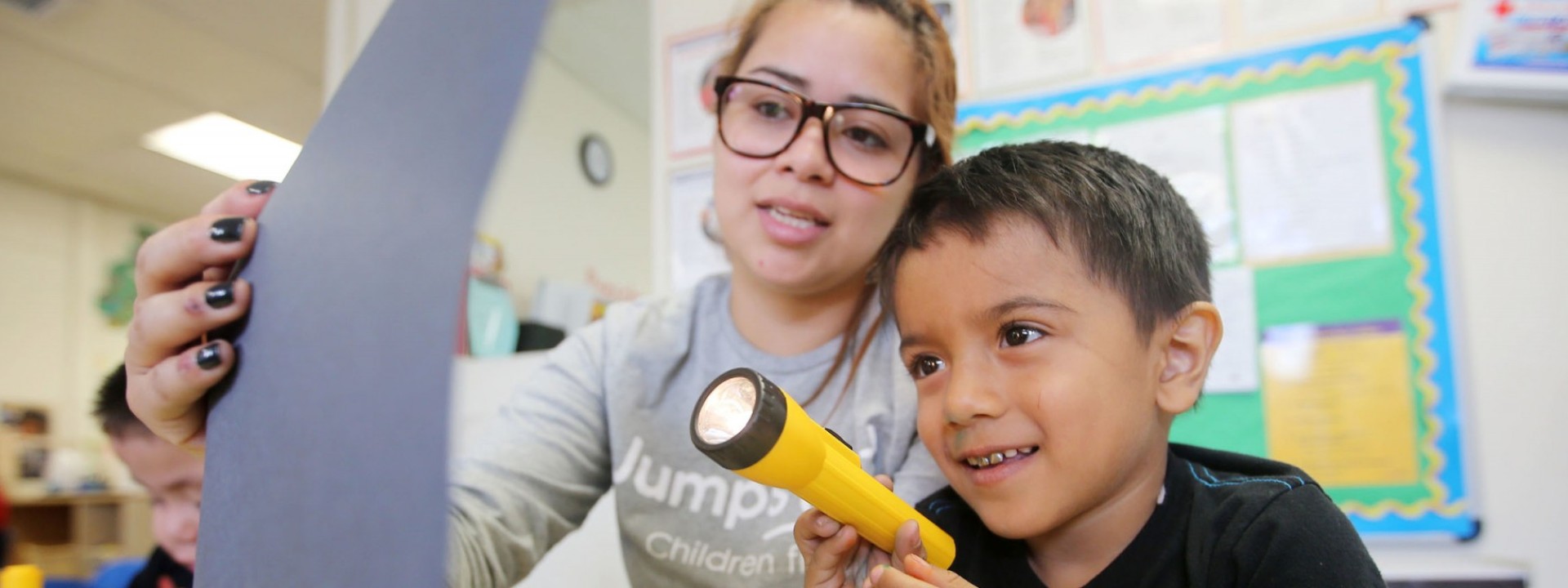 Image of a female student holding up a page for a child who is shining a flashlight on it. 