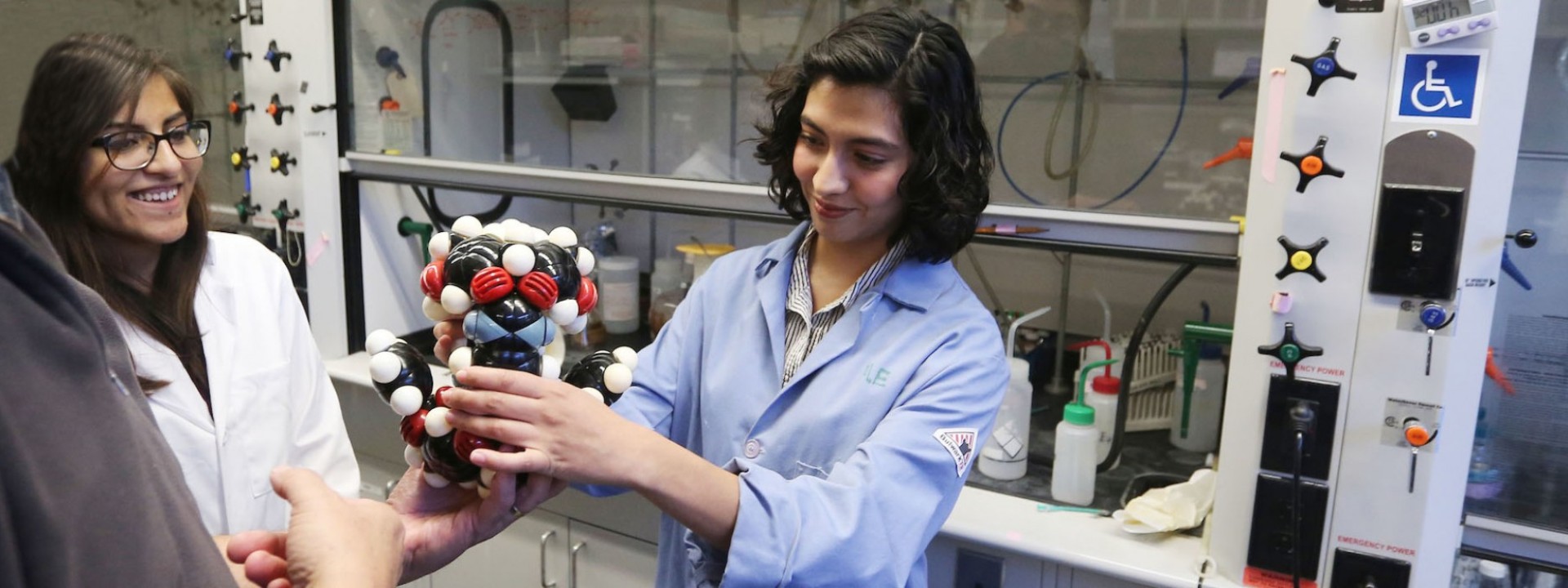 Image of a student in a blue lab coat presenting a model of an atom to a group of fellow students. 