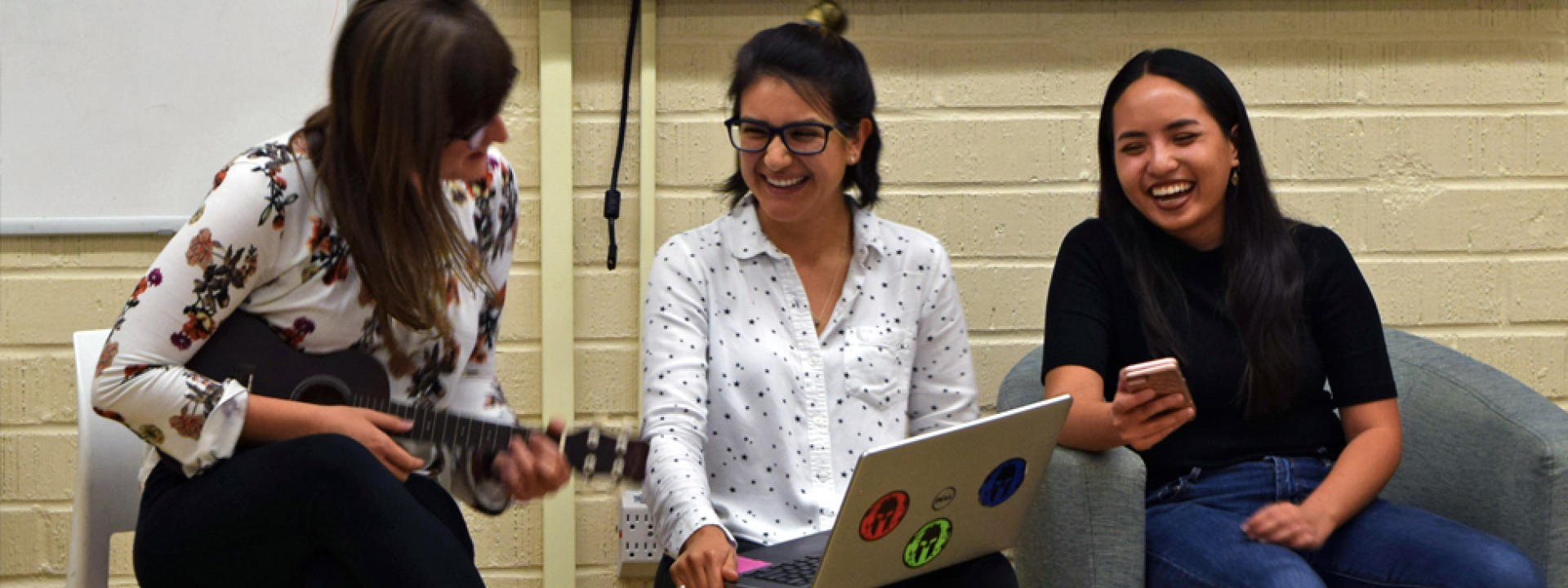 Three female students smiling, one playing a ukalele. 